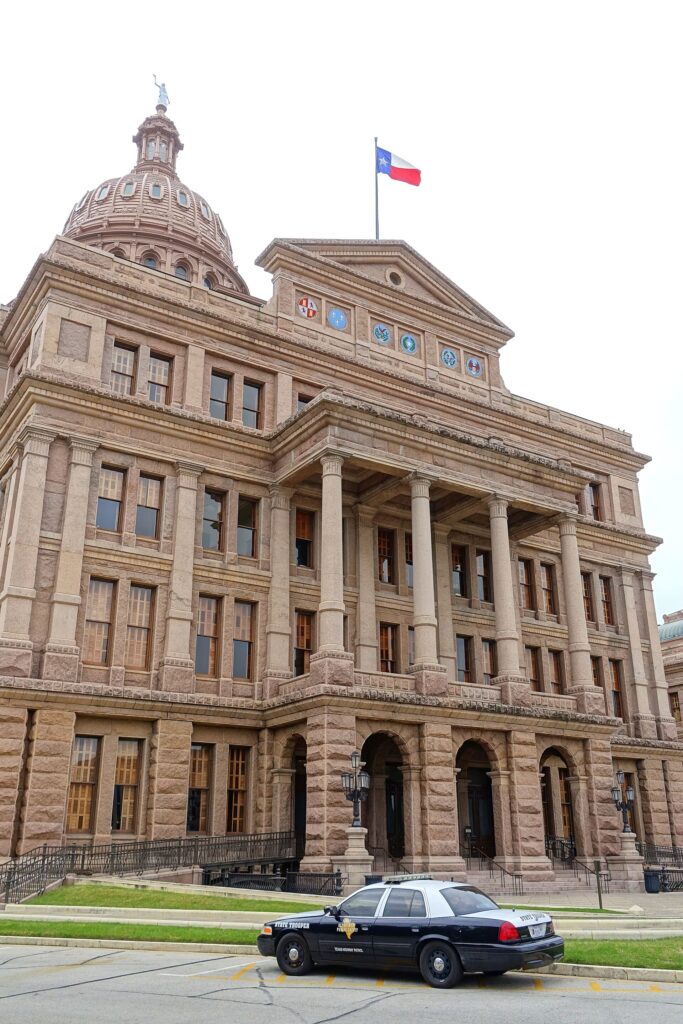 The domed Texas State Capitol in Austin, a pink granite building with a Texas flag on the roof and a police car parked out front
